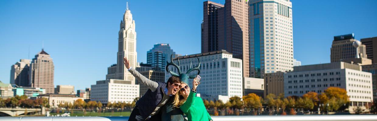 Two women posing with deer statue in front of Columbus skyline
